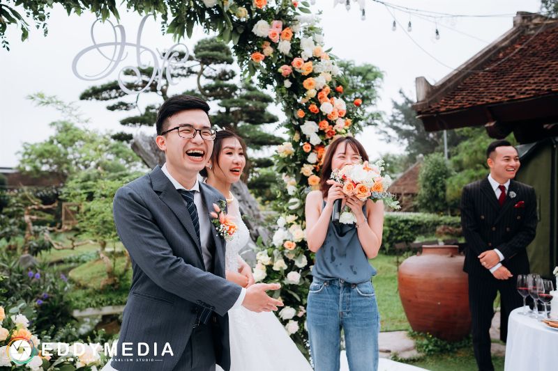 bạn cô dâu đứng cười ngại ngùng che mặt nhìn cô dâu chú rể ở cổng cưới